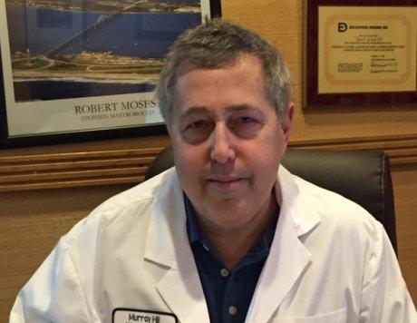 The image shows a man sitting at a desk with a plaque on the wall behind him, wearing a white lab coat and glasses, posing for a professional portrait.