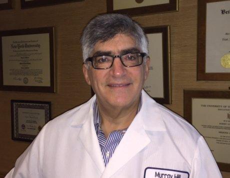 A man wearing glasses and a white lab coat stands in front of framed certificates on a wall, smiling slightly at the camera.