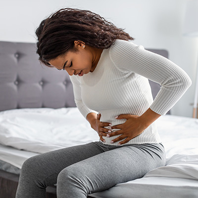 The image shows a person sitting on a bed with their hand on their stomach, suggesting discomfort or pain.