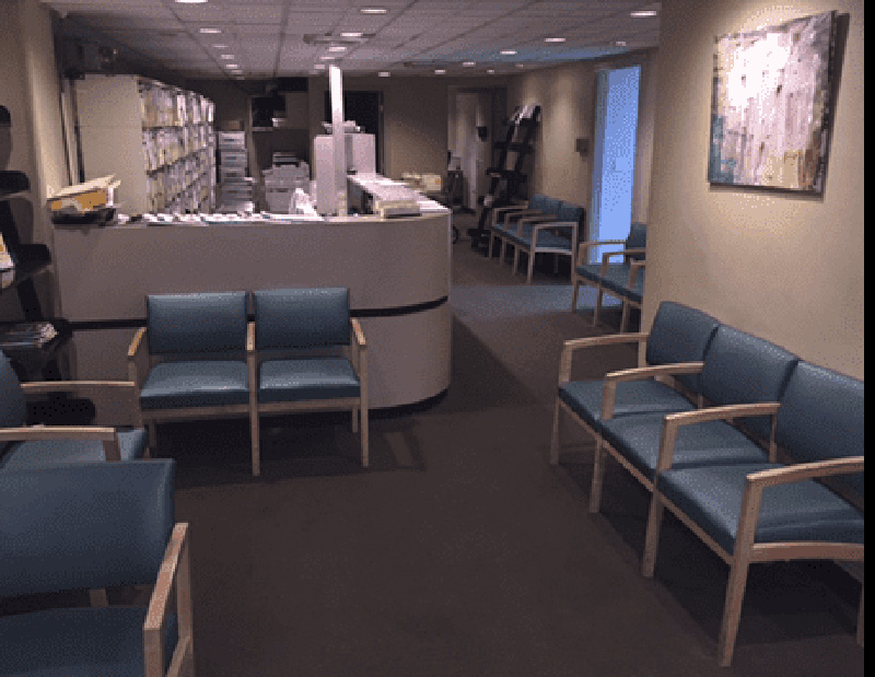 The image shows an interior space, likely a waiting area within a medical facility, featuring blue chairs arranged around a central table, with a reception desk in the background.