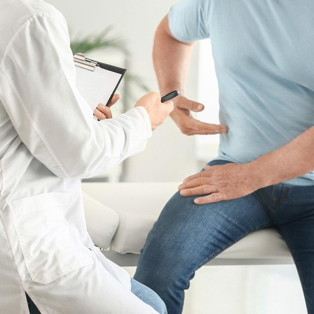 The image depicts a medical consultation between a male patient and a female doctor, with the patient seated on a chair while the doctor stands and holds a clipboard, both in an indoor setting that appears to be a medical office.