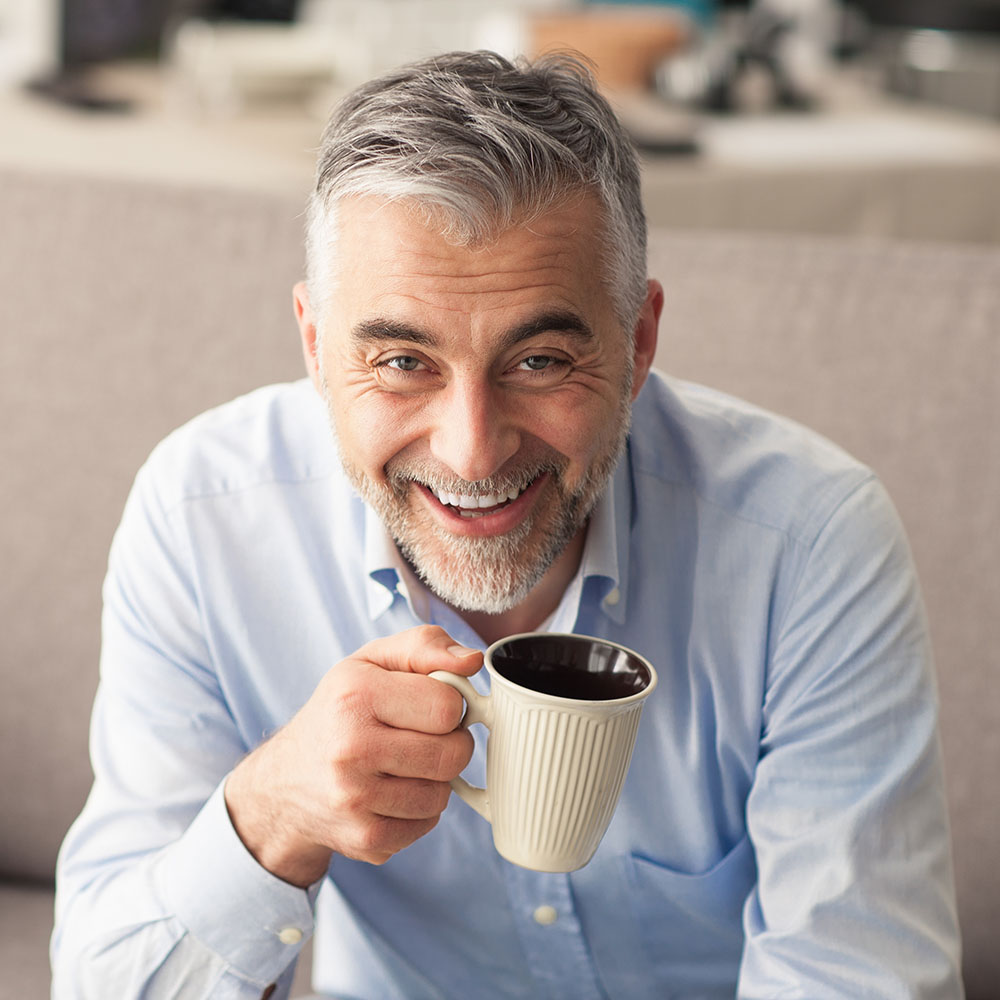 The image shows a man with gray hair, wearing a blue shirt, holding a coffee mug and smiling at the camera.